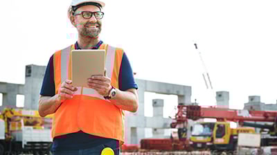 A man in an orange vest and construction hard hat holding an ipad in his hand while smiling. 